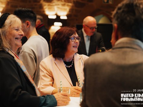In einem gemütlich beleuchteten Raum stehen mehrere Menschen an einem Stehtisch und unterhalten sich fröhlich. Zwei Frauen in der Bildmitte lächeln, während sie Getränke in der Hand halten. Im Hintergrund sind weitere Personen zu sehen, die ebenfalls in Gespräche vertieft sind. Einige Lichtakzente an der Wand schaffen eine warme Atmosphäre. Unten rechts ist ein Logo, das auf das 20-jährige Jubiläum der United Kids Foundations hinweist.