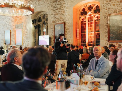 Eine festliche Veranstaltung in einem historischen Saal mit Steinwänden und großen Bogenfenstern. Gäste sitzen in eleganter Kleidung an langen, eingedeckten Tischen. Ein Kameramann filmt die Szenerie. Links hängt ein großer Kronleuchter an der Holzdecke. Am unteren Rand ist das Logo von United Kids Foundations und ein Hinweis auf das 20-jährige Jubiläum zu sehen.