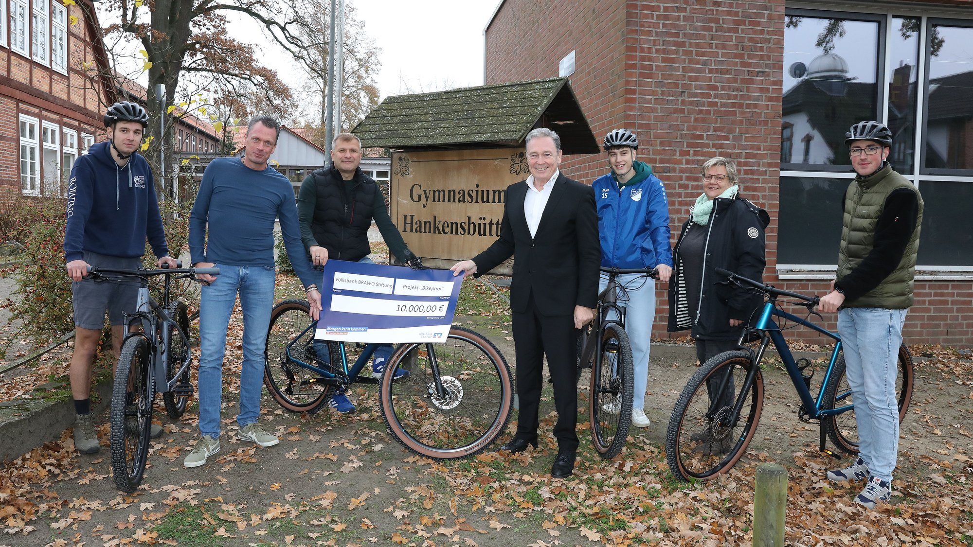 Das Bild zeigt, sieben nebeneinander stehende Personen. Jede zweite Person steht neben einem Fahrrad, die Person in der Mitte sitzt auf dem Fahrrad. Die dritte Person von links hält gemeinsam mit der Person rechts von ihm ein kleines Plakat in der Hand, dessen Aufdruck nicht zu lesen ist. Die Personengruppe steht vor zwei Gebäuden, sowie einem Schild auf dem "Gymnasium Hankensbüttel" steht. Auf dem Boden liegt vertrocknetes Laub.