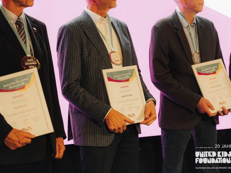 Drei Personen in Anzügen stehen nebeneinander und halten gerahmte Urkunden in ihren Händen. Jeder trägt eine Medaille um den Hals. Die Urkunden stammen von der "United Kids Foundations Ehrenmedaille". Im Hintergrund ist ein beleuchtetes Schild zu sehen, das das 20-jährige Jubiläum der United Kids Foundations feiert.