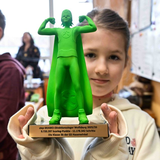 Ein Mädchen hält eine grüne Statue, die eine Superheldenfigur mit Umhang darstellt. Die Statue wird mit beiden Händen präsentiert. Darunter befindet sich eine goldene Plakette mit der Aufschrift: "step BRAWO-Direktionssieger Wolfsburg 2025/26, 9.720.297 Scoring-Punkte - 11.178.342 Schritte, Die Klasse 3b der GS Hasenwinkel." Im Hintergrund sind verschwommen andere Personen und eine Tafel erkennbar.