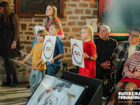 Das Bild zeigt eine Gruppe von Kindern, die Schilder mit dem Logo der "United Kids Foundations" halten. Sie stehen in einem Raum mit einer gemauerten Wand im Hintergrund. Neben den Kindern sitzen Erwachsene auf Stühlen und schauen aufmerksam in eine Richtung. Einige Personen sind elegant gekleidet. Die Szene wirkt wie eine feierliche Veranstaltung. Im Vordergrund ist unscharf ein Bildschirmrand zu sehen. In der rechten unteren Ecke befindet sich ein Logo mit der Aufschrift "20 Jahre United Kids Foundations, das Kindernetzwerk der Brawo Group".
