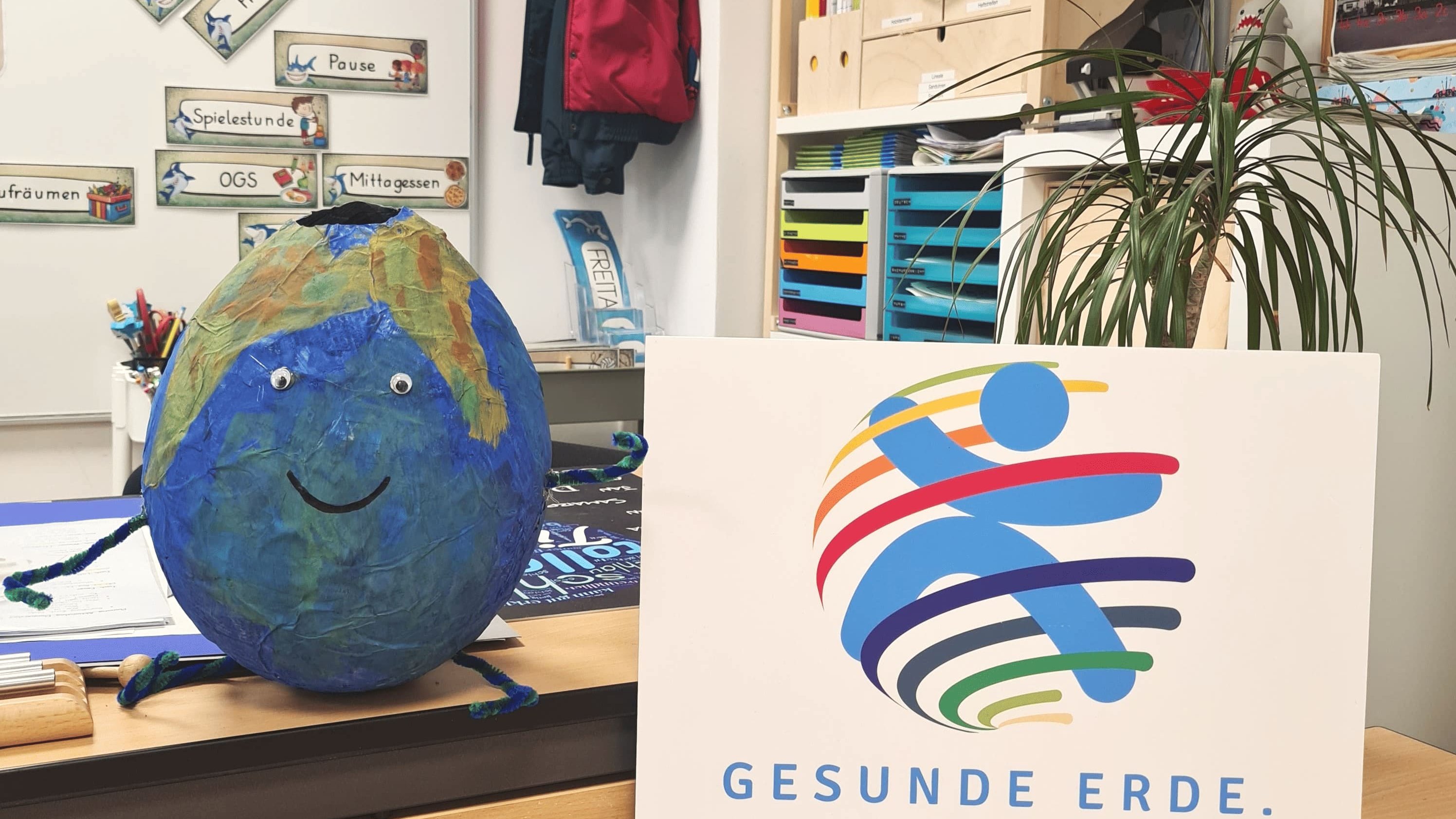 In einem Klassenraum steht auf einem Tisch eine gebastelte, lachende Erdkugel mit Augen und Armen aus Pfeifenreinigern. Daneben befindet sich ein Schild mit einem bunten Logo und dem Text "GESUNDE ERDE. GESUNDE KINDER". Im Hintergrund sind eine Pflanze, Regale und eine Pinnwand zu sehen. An der Wand hängen kleine Schilder mit verschiedenen Aufschriften wie "Hofpause" und "Spielestunde".