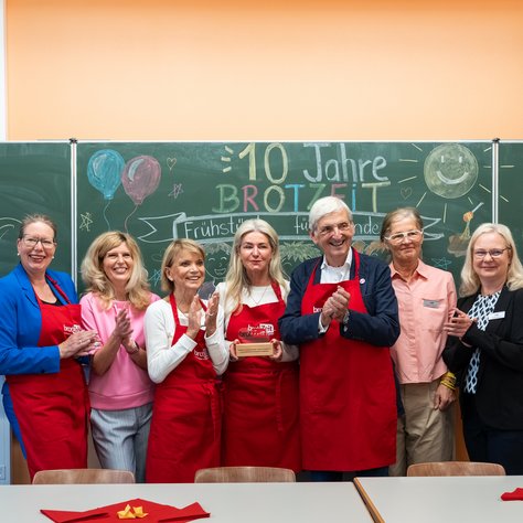 Eine Gruppe von zehn Menschen steht lächelnd in einem Raum vor einer Tafel, auf der "10 Jahre brotZeit" mit bunter Kreide geschrieben steht. Einige der Personen tragen rote Schürzen mit dem Logo von "brotZeit". Die Tafel ist mit Zeichnungen von Ballons, Sternen und einer Sonne geschmückt. Alle Personen klatschen und scheinen eine Feier zu genießen. Vor ihnen sind auf dem Tisch rote Servietten ausgebreitet.