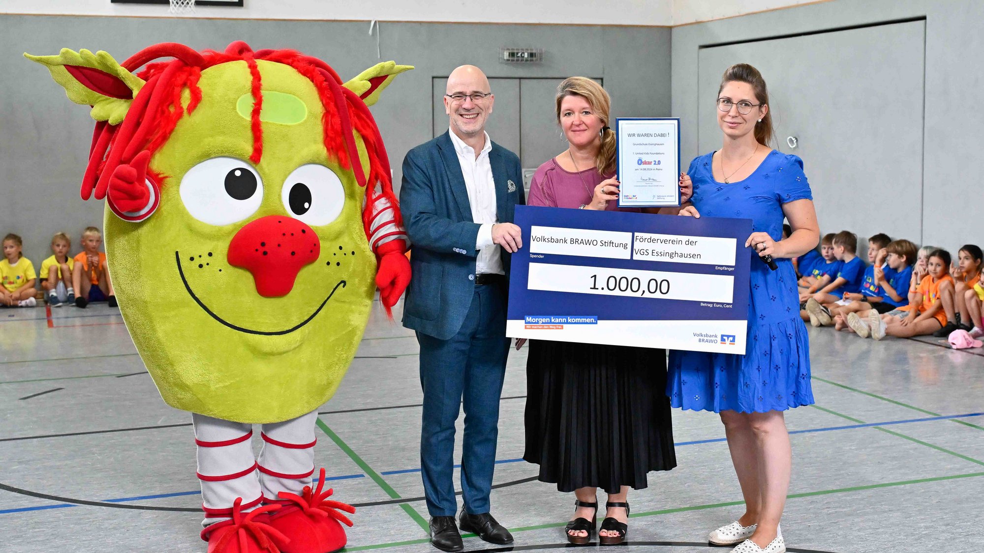 Drei Personen halten einen großen symbolischen Scheck über 1000 Euro, neben ihnen steht eine Person in einem großen gelben Maskottchenkostüm mit roten Haaren, im Hintergrund sitzen Kinder in Sportkleidung auf dem Boden einer Turnhalle.