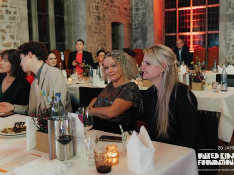 Eine Gruppe von Menschen sitzt an einem festlich gedeckten Tisch in einem Raum mit Steinmauern. Sie unterhalten sich und scheinen aufmerksam den Ereignissen zu folgen. Im Vordergrund stehen Wein- und Wassergläser sowie ein Blumenarrangement auf dem Tisch. Im Hintergrund ist schwaches, warmes Licht und eine große Fensterfront zu sehen. In der unteren rechten Ecke befindet sich das Logo der "United Kids Foundations" anlässlich ihres 20. Jubiläums.