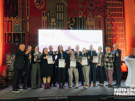 Eine Gruppe von Menschen steht auf einer Bühne vor einem kunstvoll gestalteten Wandteppich. Sie halten Urkunden und Blumensträuße in den Händen und zeigen Daumen hoch in Richtung Kamera. Die Stimmung ist feierlich. Im Hintergrund ist ein beleuchtetes Display mit der Aufschrift „20 Jahre United Kids Foundations“ zu sehen. Auf der rechten Seite steht ein kleiner weißer Stehtisch mit Blumen.