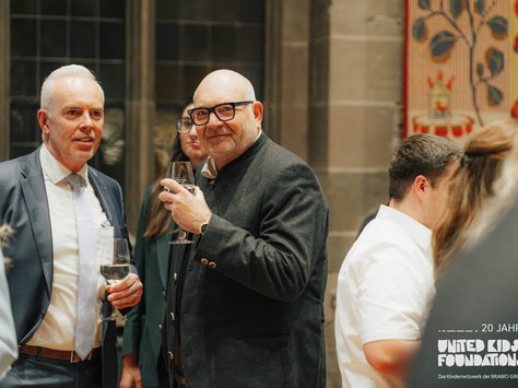 Eine Gruppe von Menschen steht bei einer Veranstaltung in einem eleganten Raum. Zwei Männer im Vordergrund, einer mit grauem Haar im Anzug und einer mit Brille und Glatze im dunklen Jackett, halten Weingläser. Im Hintergrund sind weitere Personen und ein dekorativer Wandteppich zu sehen. In der Ecke unten rechts befindet sich das Logo von "United Kids Foundations" mit dem Hinweis auf ein 20-jähriges Jubiläum.