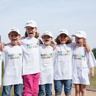 Auf dem Bild sind sechs Kinder zu sehen, die fröhlich zusammen stehen. Sie tragen weiße T-Shirts und Basecaps mit farbigem Aufdruck "United Kids Foundation". Die Kinder haben die Arme um die Schultern der anderen gelegt und lächeln breit, während einige von ihnen die Zunge herausstrecken. Im Hintergrund ist ein unscharfer Sportplatz und grünes Gras zu erkennen. Die Szenerie vermittelt eine heitere und freundliche Stimmung.