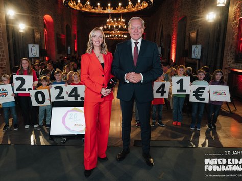 In einem festlichen Raum mit Kronleuchtern und Steinwänden stehen ein Mann im Anzug und eine Frau im roten Hosenanzug nebeneinander und lächeln in die Kamera. Sie hält ein Mikrofon. Hinter ihnen stehen Kinder je mit einem Schild, die zusammen "20.241 €" zeigen. Auf einem Monitor im Vordergrund steht "20 Jahre" und daneben ist das Logo von United Kids Foundations zu sehen. Im Hintergrund sitzen Menschen an Tischen und die Stimmung ist feierlich.
