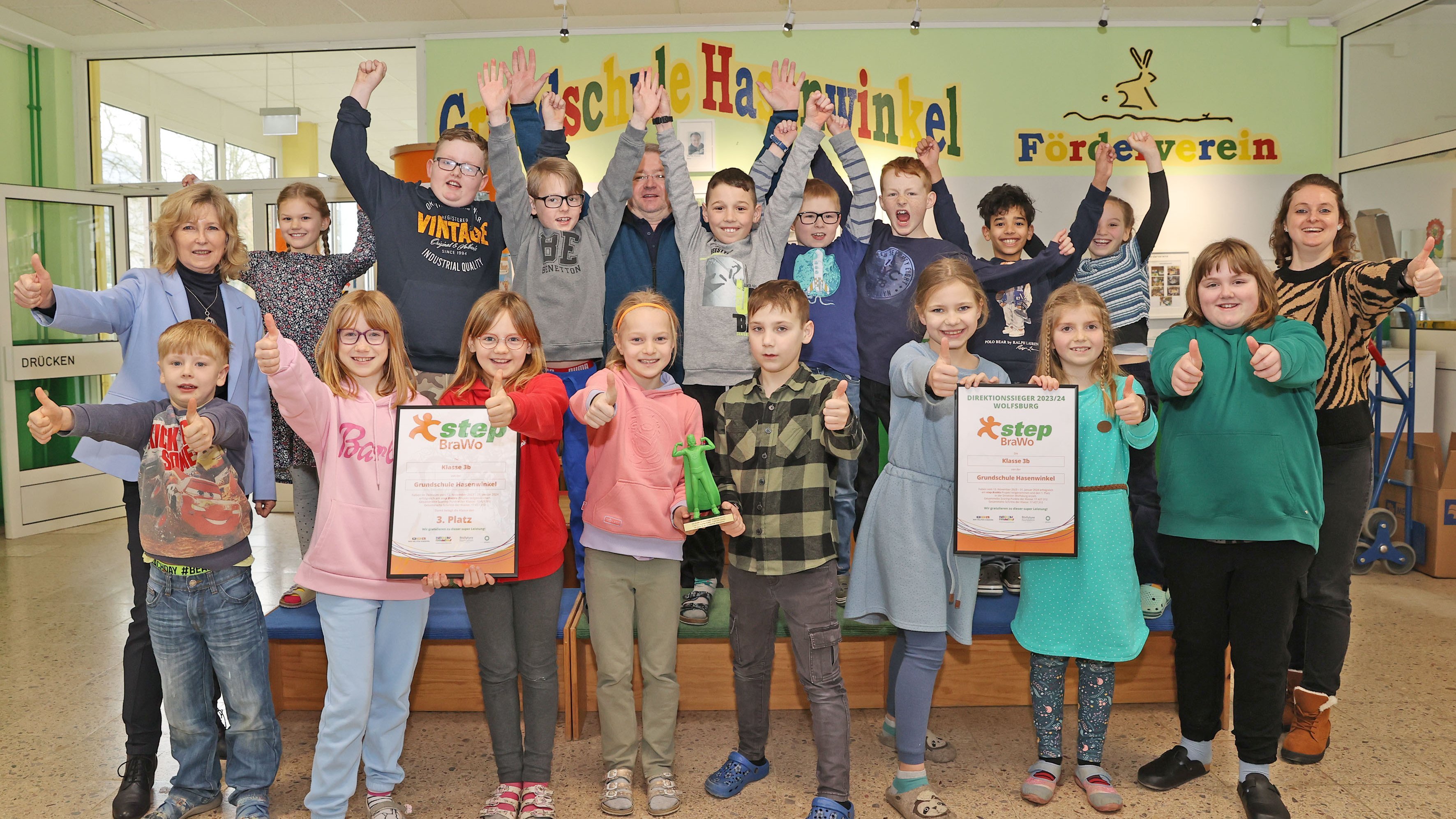 Eine Gruppe Kinder steht in zwei Reihen lächelnd vor einer Wand mit der bunten Aufschrift "Grundschule Hasenwinkel". Sie strecken ihren Arm nach vorne und zeigen mit dem Daumen nach oben.