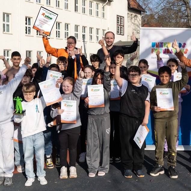 Eine Gruppe von Kindern und Erwachsenen posiert freudig mit Zertifikaten und einem gerahmten Dokument. Sie stehen vor einem Gebäude im Freien und heben Daumen hoch in die Kamera. Ein Banner mit dem Text "United Kids Foundations" ist im Hintergrund sichtbar. Die Kinder tragen bequeme Freizeitkleidung und wirken stolz und begeistert.