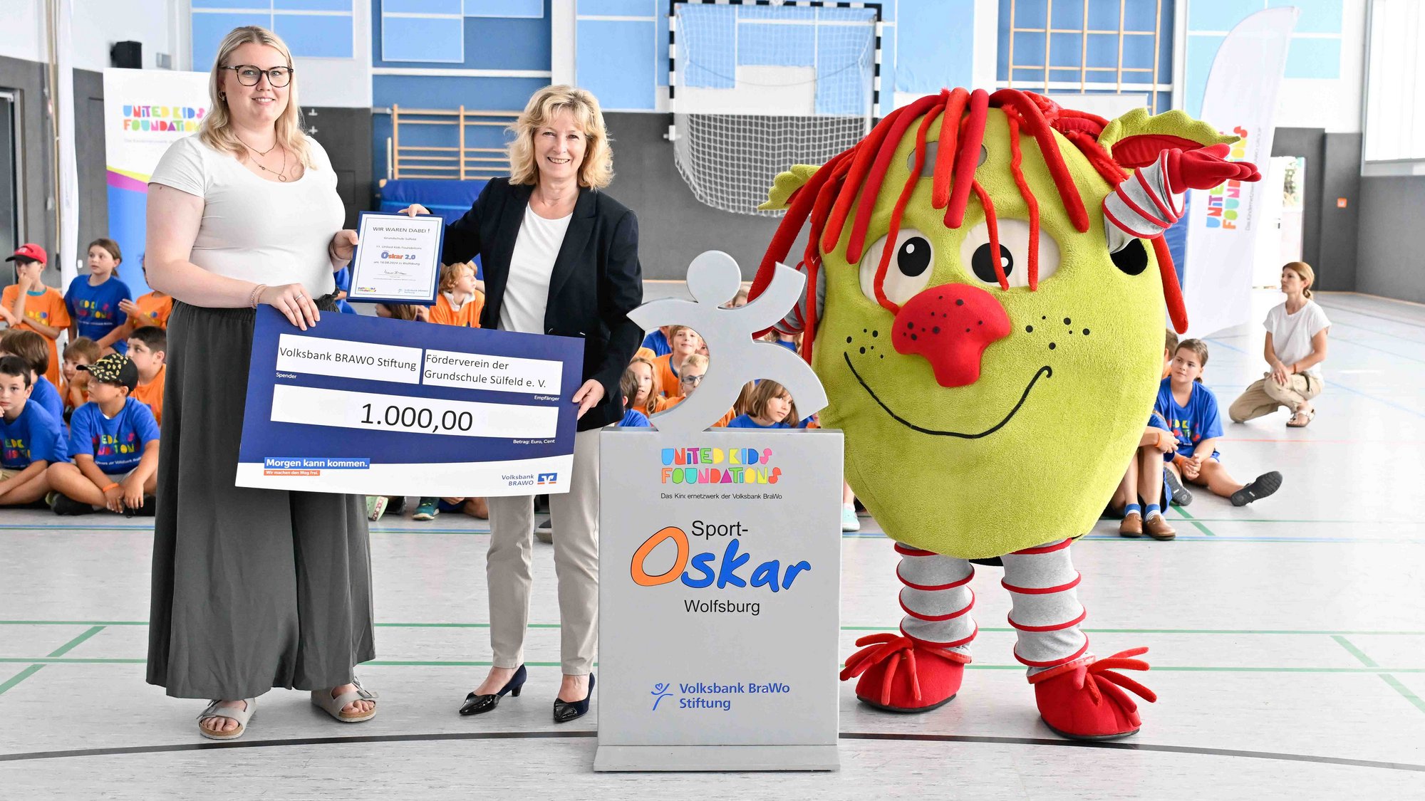Claudia Kayser, Leiterin der Direktion Wolfsburg bei der Volksbank BRAWO und Botschafterin von United Kids Foundations, übergab den symbolischen Spendenscheck an Schulleiterin Linda Bausewein (links). Foto: Volksbank BRAWO/Isabell Massel Auf dem Bild stehen zwei Personen mit langen blonden Haaren, die gemeinsam einen Spendenscheck und eine Urkunde in der Hand halten. Auf dem Spendenscheck steht groß die Zahl 1.000,00. Neben den Personen steht ein Maskottchen, einer grünen Kartoffel mit Augen, einer großen roten Nase, einem schwarzen Stich als Mund, der lächelt und schwarzen Sommersprossen über die Nase. Das Maskottchen hat große Harre und streckt die Arme in die Luft. Es trägt rote Schuhe, rote Handschuhe, einen weiß-rot gestreiften Ganzkörperanzug unter seiner Kartoffelform. Zwischen den Personen und dem Maskottchen steht ein grauer Aufsteller mit einer grauen Strichmännchen-Figur. Auf dem Aufsteller steht "United Kids Foundations Sport-Oskar Wolfsburg Volksbank BraWo Stiftung".