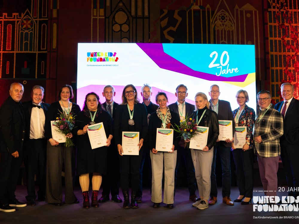 Eine Gruppe von zwölf Personen steht auf einer Bühne vor einem großen Bildschirm, der "20 Jahre United Kids Foundations" anzeigt. Einige Personen halten Blumensträuße und Urkunden. Die Stimmung ist feierlich und die Personen lächeln oder blicken stolz in die Kamera. Der Hintergrund zeigt ein stilisiertes Wandmuster und rote Beleuchtung.