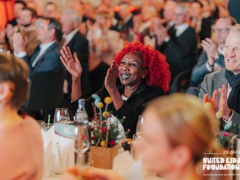 Eine Gruppe von Menschen sitzt in einem Veranstaltungsraum und applaudiert begeistert. Im Vordergrund sitzt eine lächelnde Frau mit auffälliger roter Lockenfrisur. Auf den Tischen stehen dekorative Blumenarrangements und Gläser. Die Atmosphäre wirkt festlich und lebhaft. In der rechten unteren Ecke ist das Logo der „United Kids Foundations“ zu sehen.