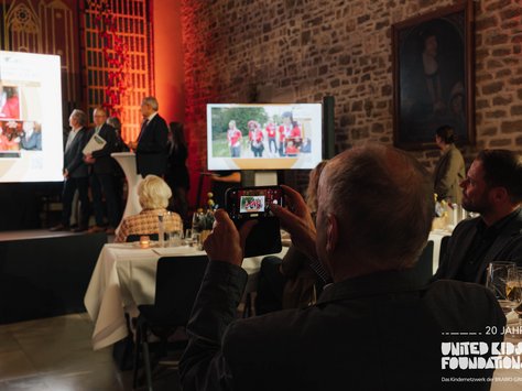 Eine Veranstaltung in einem Saal mit Ziegelwänden und gedämpfter Beleuchtung. Im Vordergrund hält eine Person ein Smartphone und fotografiert den Raum. Auf der Bühne stehen mehrere Personen neben einem Rednerpult, wobei auf einem großen Bildschirm Präsentationen mit Bildern von Menschen in roten T-Shirts gezeigt werden. Gäste sitzen an Tischen mit weißem Tischtuch, Essgeschirr und Getränken. An der Wand hängt ein großes Gemälde. Im unteren rechten Bildbereich ist das Logo von "United Kids Foundations" zusammen mit dem Text "20 Jahre" zu sehen.