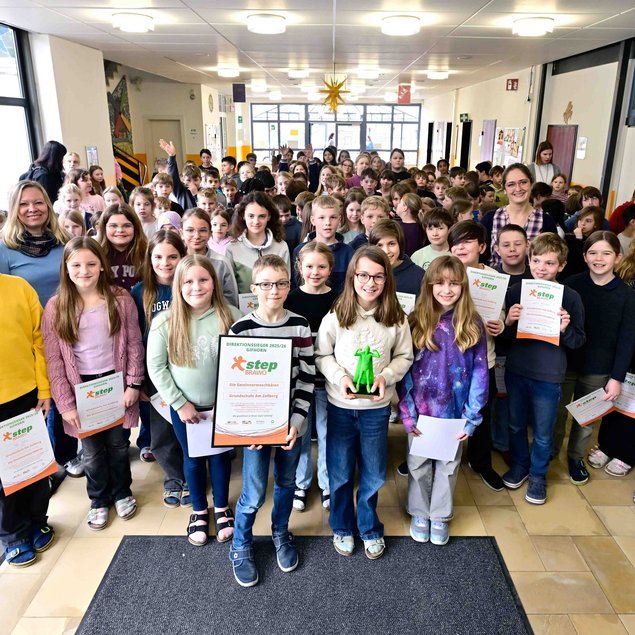 In einem hellen Flur stehen zahlreiche Kinder und einige Erwachsene zusammen. Die vorderste Reihe umfasst lächelnde Mädchen und Jungen, die Urkunden und eine grüne Statue in Händen halten. Im Hintergrund sind weitere Kinder zu sehen, die ebenfalls Urkunden halten. Die Stimmung wirkt fröhlich und feierlich.
