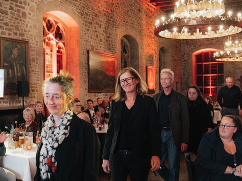 In einem festlichen Raum mit Steinwänden und großen Fenstern gehen mehrere Personen in eleganter Kleidung durch den Saal. Im Hintergrund sitzen Gäste an Tischen, die dekorativ eingedeckt sind. An der Decke hängen große Kronleuchter, die den Raum in warmem Licht erstrahlen lassen. Rechts unten im Bild ist ein Logo mit dem Text „20 Jahre United Kids Foundations“ zu sehen.