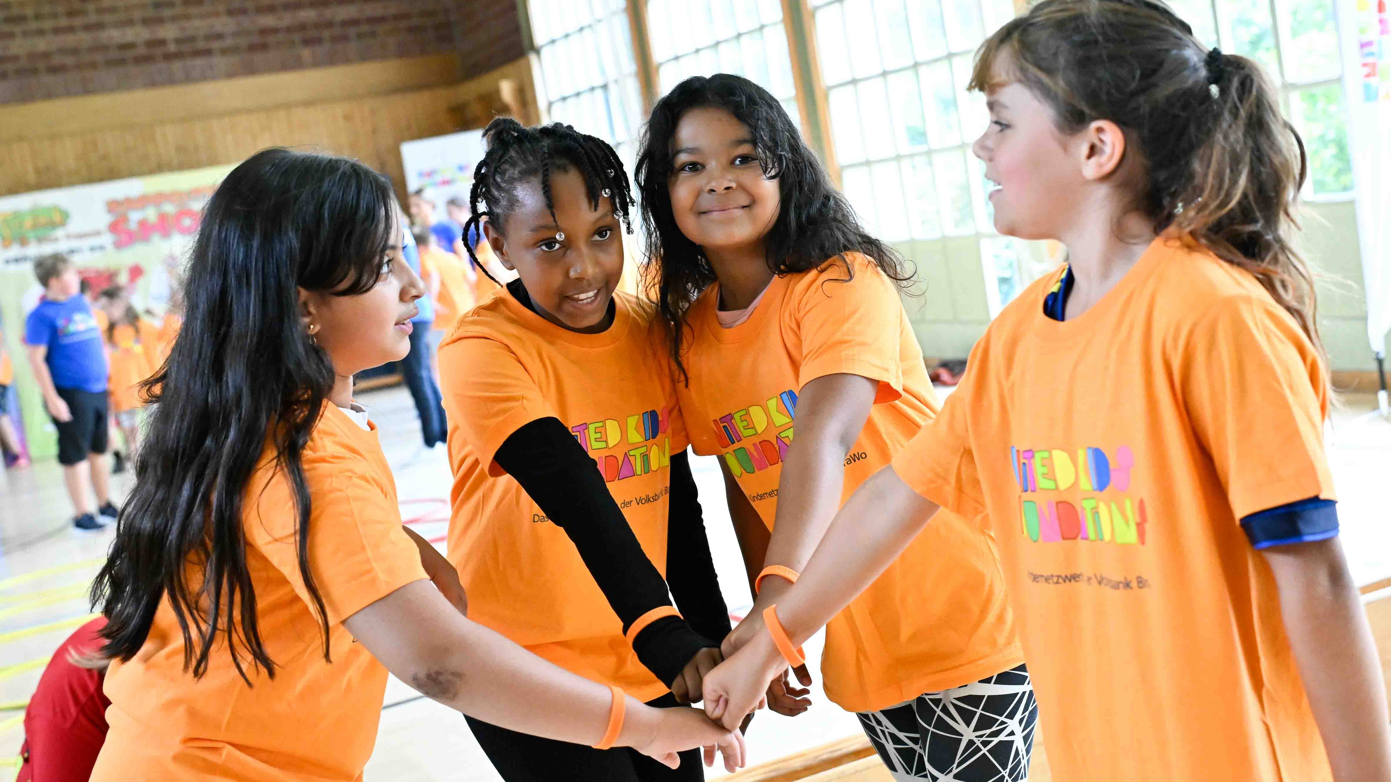 Vier Kinder stehen in einem Kreis und tragen orangefarbene T-Shirts mit buntem Schriftzug. Sie legen die Hände übereinander in die Mitte und scheinen sich auf eine gemeinsame Aktivität vorzubereiten. Im Hintergrund sieht man weitere Kinder in der Sporthalle. Die Stimmung wirkt fröhlich und kameradschaftlich.