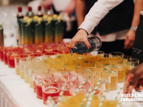 Eine Person gießt Wasser aus einer Flasche in ein Glas. Auf einem Tisch stehen zahlreiche Gläser mit roten und gelben Getränken, jeweils mit Zitronenscheiben garniert. Im Hintergrund sind mehrere Flaschen mit unterschiedlichen Getränken zu sehen. Auf dem Bild befindet sich ein Logo, das auf eine Veranstaltung der "United Kids Foundations" hinweist.