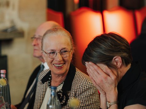 Zwei Frauen sitzen an einem Tisch in einem festlich beleuchteten Raum. Die ältere Frau lächelt und trägt eine Brille und ein kariertes Jackett. Die jüngere Frau neben ihr lacht und hält sich die Hände vors Gesicht. Vor ihnen stehen Wasserflaschen und Blumendekorationen auf dem Tisch. Im Hintergrund sind verschwommen einige Stühle und ein rot illuminierter Bereich zu sehen. Unten rechts im Bild befindet sich das Logo von „United Kids Foundations“ und ein Hinweis auf das 20-jährige Jubiläum.