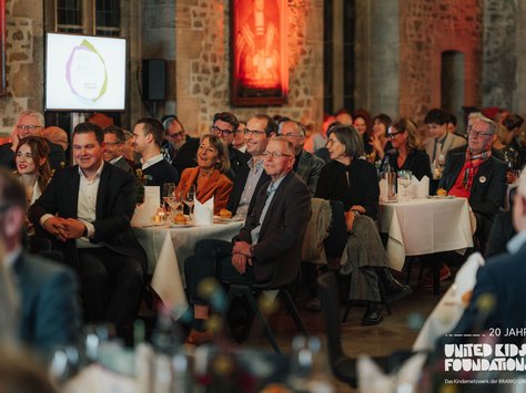 Eine Gruppe von Menschen sitzt an festlich gedeckten Tischen in einem großen Raum mit Backsteinwänden. Sie wirken aufmerksam und lächeln, als würden sie einer Präsentation folgen. Im Hintergrund hängt ein Gemälde, und auf einem Bildschirm steht "20 Jahre". Auf einem Tisch stehen Weingläser und brennende Kerzen. Die Atmosphäre ist gemütlich und feierlich. In der Ecke ist der Schriftzug "United Kids Foundations" zu sehen.