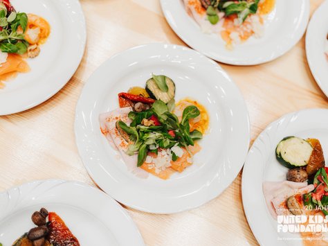 Das Bild zeigt mehrere weiße Teller auf einem Holztisch, die jeweils mit einer kunstvoll angerichteten Vorspeise dekoriert sind. Die Teller enthalten verschiedene Zutaten wie dünn geschnittenen Lachs, Salatblätter, Zucchini, Paprika und eventuell einige Pilze. Auf einem der Teller ist das Logo der "United Kids Foundation" abgebildet.