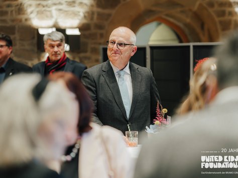 Bei einer Veranstaltung steht ein Mann mittleren Alters in einem grauen Anzug und mit Brille an einem Tisch, auf dem ein Glas Getränk und Dekorationen stehen. Im Hintergrund sind weitere Personen in einem Raum mit Steinmauern zu sehen. Es handelt sich um eine Feier oder ein Treffen im Rahmen des 20-jährigen Jubiläums der "United Kids Foundations".