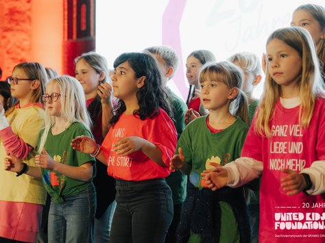 Eine Gruppe von Kindern steht dicht beisammen und singt oder führt eine Art von Performance auf. Sie tragen bunte T-Shirts in verschiedenen Farben wie grün, rot und pink, mit teilweise bedruckten Slogans und Grafiken. Die Umgebung ist warm beleuchtet, und im Hintergrund ist eine große leuchtende Fläche zu sehen, die als Projektionsfläche dient. Die Kinder wirken konzentriert und fröhlich. In der Ecke des Bildes befindet sich ein Logo mit der Aufschrift "20 Jahre United Kids Foundations".
