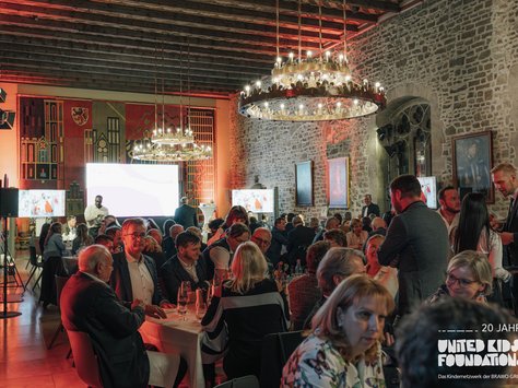 Das Bild zeigt eine festliche Veranstaltung in einem historischen Saal mit rustikalen Steinwänden. An mehreren runden Tischen sind zahlreiche Gäste versammelt, die in Gespräche vertieft sind. Zwei große, dekorative Kronleuchter beleuchten den Raum, der mit kunstvollen Wandteppichen und Gemälden geschmückt ist. Im Hintergrund sind mehrere Bildschirme zu sehen, und die Beleuchtung im Raum ist in warmen Rot- und Orangetönen gehalten. In der rechten unteren Ecke des Bildes ist das Logo der "United Kids Foundations" mit einem Hinweis auf das 20-jährige Jubiläum zu sehen.