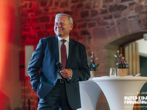 Ein älterer Mann in einem Anzug steht lächelnd auf einer Bühne. Er hält ein Mikrofon in der Hand. Der Hintergrund ist aus rotem Backstein und weich beleuchtet. Neben ihm ist ein Stehtisch mit einem Glas Wasser und einer Blumendekoration zu sehen. In der Ecke ist das Logo von United Kids Foundations mit dem Hinweis "20 Jahre" zu erkennen.