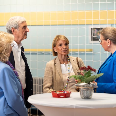 Drei Frauen und ein Mann stehen um einen Stehtisch und unterhalten sich in einem Raum mit hellblauen Wänden und gelben Akzenten. Auf dem Tisch steht eine Pflanze in einem dekorativen Übertopf und ein kleines Körbchen mit Süßigkeiten. Im Hintergrund sind gerahmte Bilder an der Wand zu sehen. Alle Personen sind formell gekleidet.
