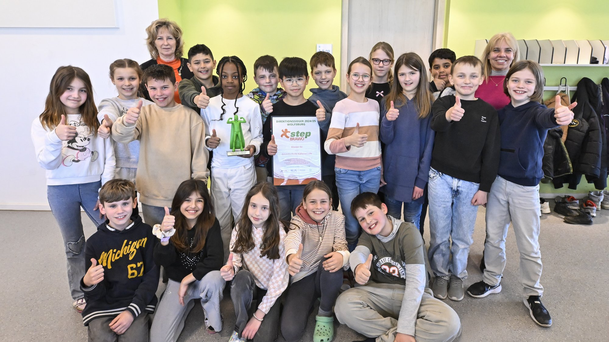 Auf dem Bild sieht man eine Gruppe von Kindern, die in die Kamera lächeln. Einige von Ihnen zeigen mit dem Daumen nach oben und zwei Kinder halten jeweils den step BRAWO-Pokal und die Urkunde in der Hand. Hinter den Kindern steht links eine in orange gekleidete blonde Frau und rechts eine in pink gekleidete blonde Frau.