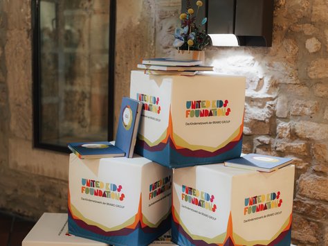 Das Bild zeigt mehrere Würfel mit dem Logo "United Kids Foundations" vor einer Steinwand. Auf den Würfeln liegen Bücher und obenauf befindet sich eine kleine Vase mit bunten Trockenblumen. Neben den Würfeln steht "20 Jahre United Kids Foundations - Das Kindernetzwerk der BraWo Group" geschrieben. Im Hintergrund ist die spiegelnde Glastür eines Raums zu sehen, der rustikal gestaltet ist.