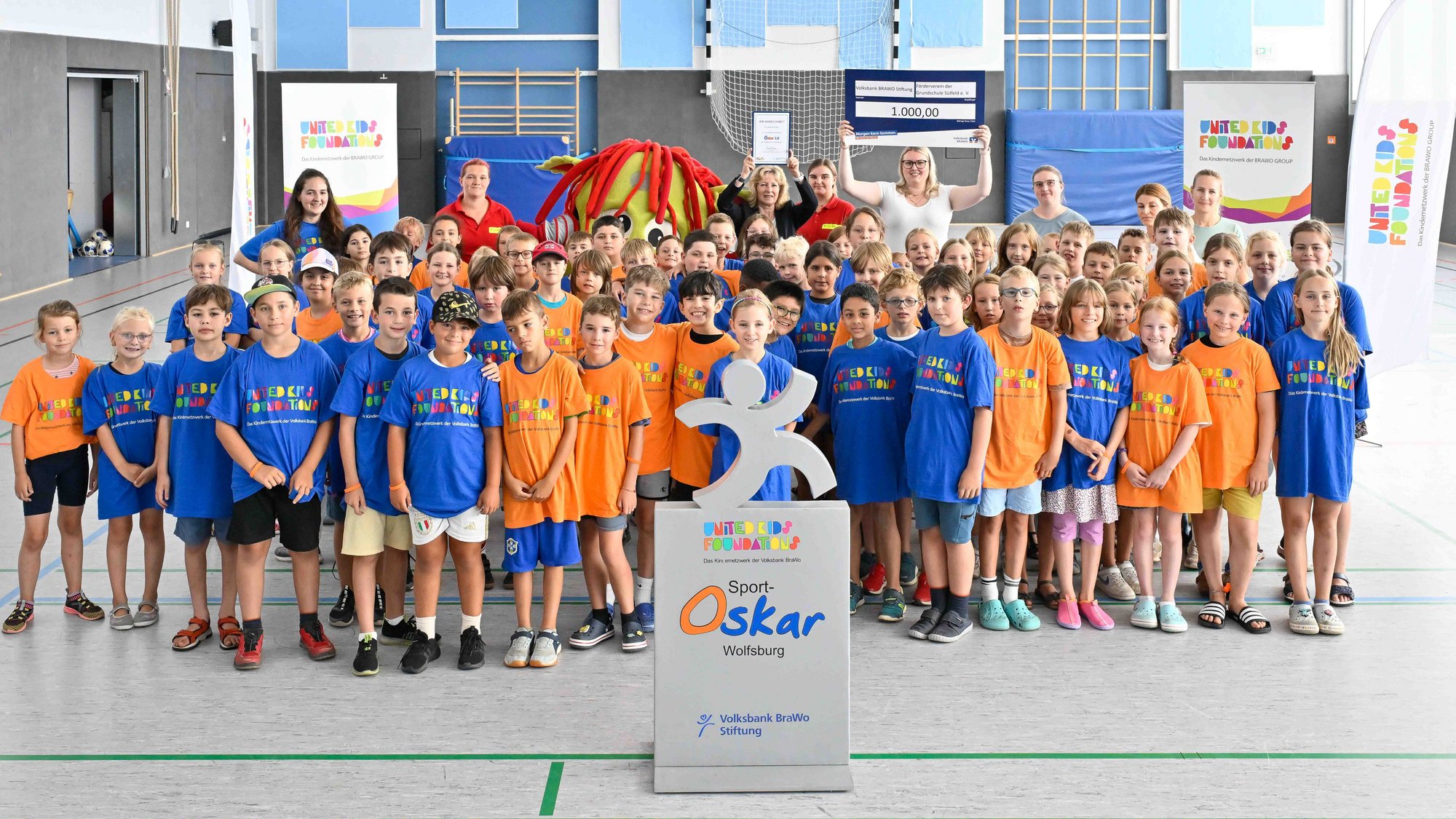 Der große Bewegungstag im Rahmen des Sport-Oskars motivierte die gesamte Grundschule Sülfeld zu einem Tag voller Spiel, Sport und Spaß. Foto: Volksbank BRAWO/Isabell Massel Auf dem Bild ist eine große Gruppe von Kindern, so wie vereinzelt Erwachsene zu sehen. Die Gruppe steht in einer Turnhalle. Vor der Gruppe steht ein kleinerer, silberner Aufsteller, auf dem eine silbernes Strichmännchen steht, der gerade einen großen Schritt zu machen scheint.