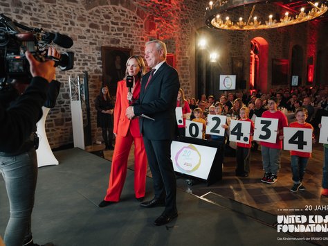 In einem großen Raum mit Steinwänden findet eine Veranstaltung statt. Vorne stehen eine Frau in einem roten Anzug und ein Mann in einem dunklen Anzug, beide lächeln und halten Mikrofone. Eine Kamera filmt sie. Daneben stehen Kinder in bunten T-Shirts, die große Zahlen auf Schildern halten. Im Hintergrund applaudieren Menschen an festlich gedeckten Tischen. Der Raum ist festlich beleuchtet mit großen Kronleuchtern. Auf einem Bildschirm ist „20 Jahre United Kids Foundations“ zu sehen.