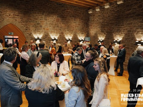 Eine Gruppe von Menschen steht in einem festlichen Raum mit Steinwänden und Holzboden. Sie unterhalten sich in kleinen Gruppen an Stehtischen. Einige Gäste genießen Getränke und Snacks. Im Hintergrund ist ein Banner zu sehen, das auf das 20-jährige Jubiläum von "United Kids Foundations" hinweist. Die Stimmung ist lebhaft und gesellig.