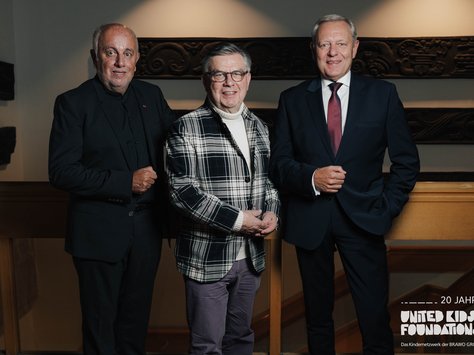 Drei Männer stehen nebeneinander in einem Innenraum. Der Mann links trägt einen schwarzen Anzug und lächelt in die Kamera. Der Mann in der Mitte hat ein kariertes Sakko an und trägt eine Brille. Der Mann rechts trägt einen dunklen Anzug mit roter Krawatte. Im Hintergrund ist eine kunstvoll geschnitzte Holzverkleidung zu sehen. In der Ecke unten rechts steht das Logo der "United Kids Foundations" mit dem Zusatz "20 Jahre".