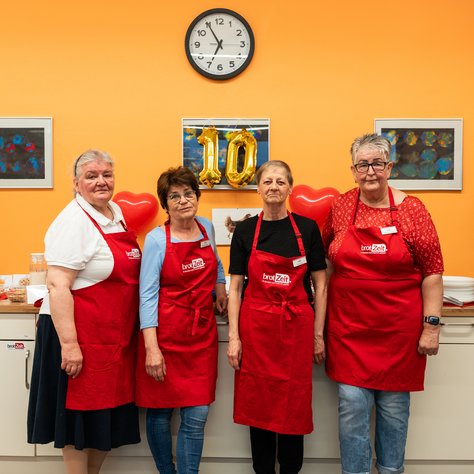 Vier Personen stehen in einer Küche vor einer orangefarbenen Wand und tragen rote Schürzen mit der Aufschrift „brotZeit“. Sie lächeln und stehen nebeneinander. Hinter ihnen sind goldene Luftballons mit der Zahl „10“ und zwei rote herzförmige Ballons zu sehen. Eine Wanduhr zeigt die Zeit an. Im Hintergrund hängen zwei Bilder an der Wand.