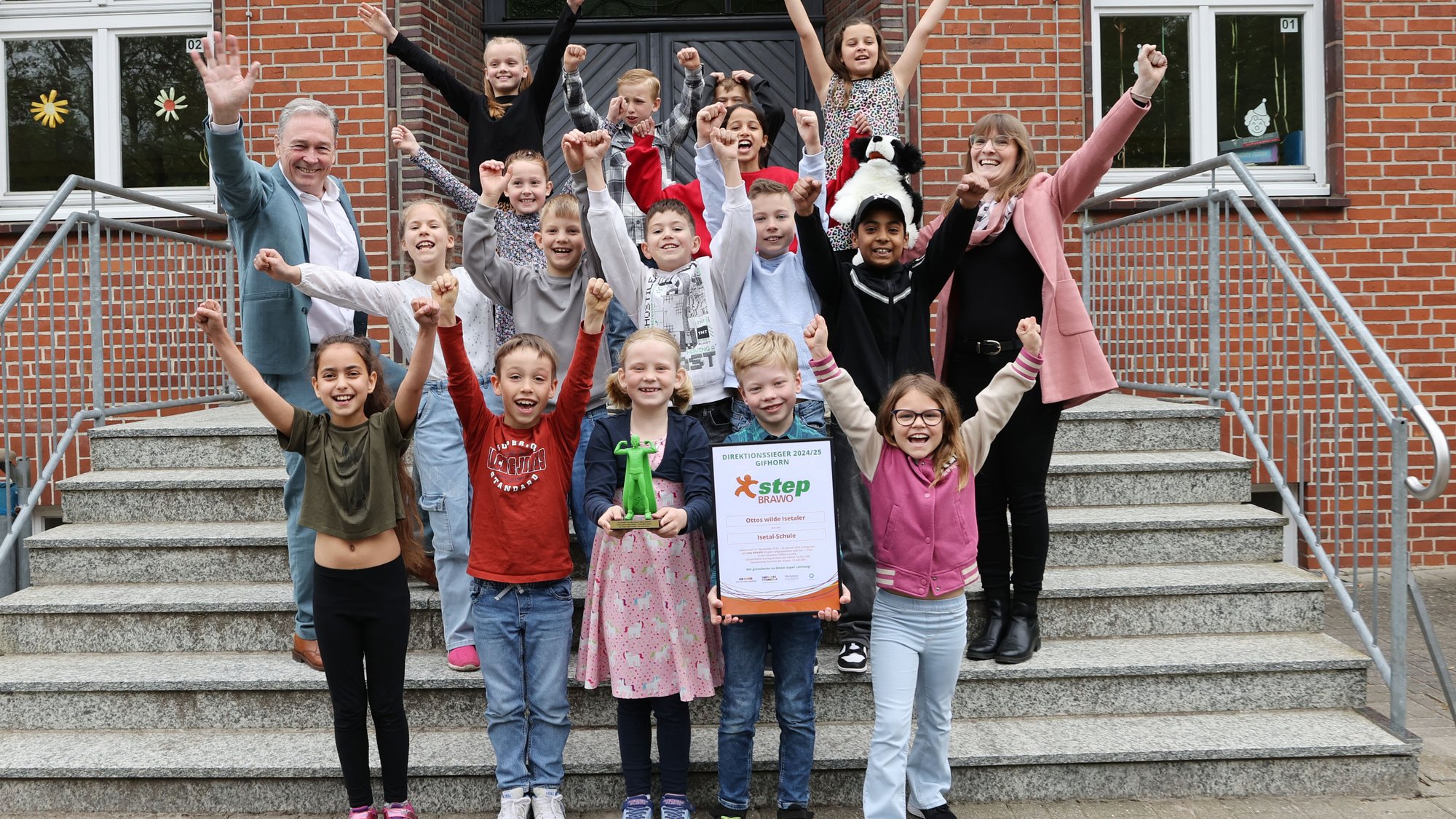 Eine fröhliche Kindergruppe steht mit zwei Erwachsenen jubelnd auf einer Treppe vor einem Schulgebäude. Ein Junge in der ersten Reihe hält stolz eine Urkunde, ein anderes Kind eine grüne Trophäe. Alle heben die Arme in die Luft und lachen, offensichtlich anlässlich einer Siegerehrung oder eines besonderen Erfolges.