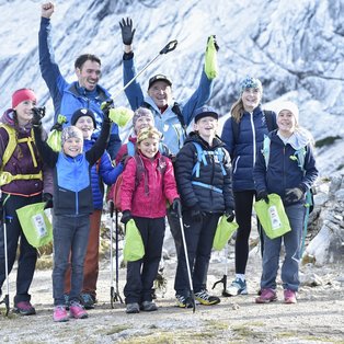 Eine Gruppe von glücklichen Menschen unterschiedlicher Altersgruppen steht zusammen auf einem Bergpfad vor einer schneebedeckten Berglandschaft. Sie tragen Outdoor-Kleidung und Wanderstöcke, einige heben triumphierend ihre Arme in die Luft und lächeln in die Kamera. Sie halten grüne Taschen in den Händen. Die Umgebung ist felsig und karg, mit einigen Schneeflecken auf dem Boden.