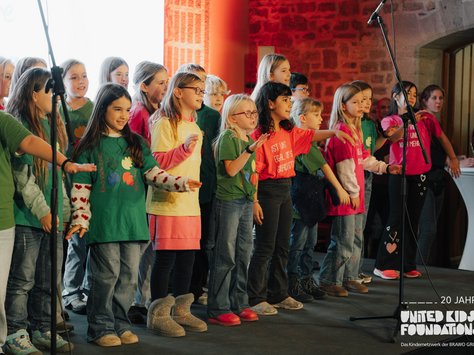 Eine Gruppe von Kindern steht auf einer Bühne und singt. Sie tragen bunte T-Shirts und Jeans, einige mit Aufschriften. Im Vordergrund sind Mikrofone aufgestellt. Der Hintergrund ist in roten und weißen Farbtönen gehalten, und auf der rechten Seite sind einige Erwachsene zu sehen, die zuschauen. Unten rechts im Bild ist das Logo der "United Kids Foundations" mit dem Text "20 Jahre".