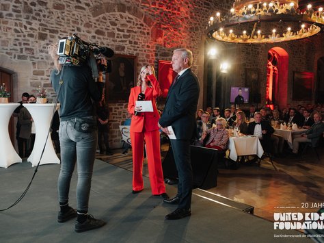 In einem festlichen, historischen Raum mit Steinwänden und Porträts an den Wänden steht eine Frau in einem leuchtend roten Anzug neben einem Mann in dunklem Anzug. Beide halten Mikrofone und sprechen zu einem Publikum. Unweit von ihnen filmt ein Kameramann die Szene. Die Zuschauenden sitzen an festlich gedeckten Tischen, und im Hintergrund ist ein großer Kronleuchter sichtbar. Text im Bild: "20 Jahre United Kids Foundations, Das Kindernetzwerk der BraWo Group".