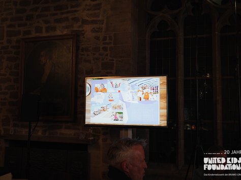 Das Bild zeigt den Innenraum eines Gebäudes mit Steinwänden. Auf der linken Seite ist ein dunkles Gemälde zu sehen. Rechts davon befindet sich ein beleuchteter Bildschirm, der eine bunte Präsentation mit mehreren Bildern von lächelnden Kindern und Zahlen anzeigt. Die rechten Seite des Bildes besteht aus zwei hohen, gotischen Fenstern. Im Vordergrund unten ist ein Teil eines Kopfes einer Person sichtbar. Unten rechts steht der Text: "20 JAHRE UNITED KIDS FOUNDATIONS Das Kindernetzwerk der BRAWO GROUP."
