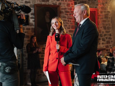 Auf dem Bild sieht man eine Frau im roten Anzug, die ein Mikrofon mit dem Logo von "ntv" hält. Sie steht neben einem älteren Mann in einem dunklen Anzug. Im Hintergrund ist eine Kamera und ein Kameramann zu sehen, ebenso ein gemauertes Zimmer mit einem Gemälde an der Wand. Einige Personen im Hintergrund klatschen. Unten rechts befindet sich ein Logo mit der Aufschrift "20 Jahre United Kids Foundations, das Kindernetzwerk der BRAVO GROUP".
