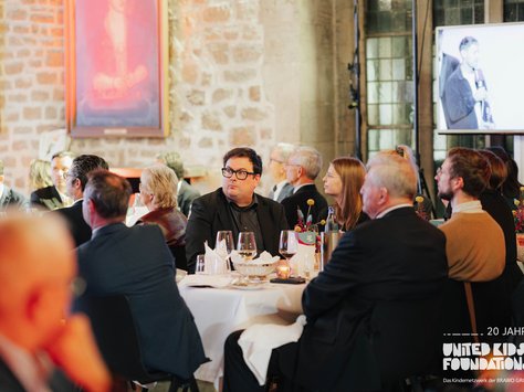 In einem stilvollen Raum mit Steinwänden und großen Porträts sitzen elegant gekleidete Menschen an gedeckten Tischen. Auf einem Bildschirm im Hintergrund ist eine Live-Übertragung von Sprechern zu sehen. Auf den Tischen stehen Weingläser und Blumenarrangements. Ein Logo mit der Aufschrift „20 Jahre United Kids Foundations – Das Kinder-Netzwerk der BRAVO Group“ ist eingeblendet. Die Anwesenden schauen konzentriert zur Bühne.