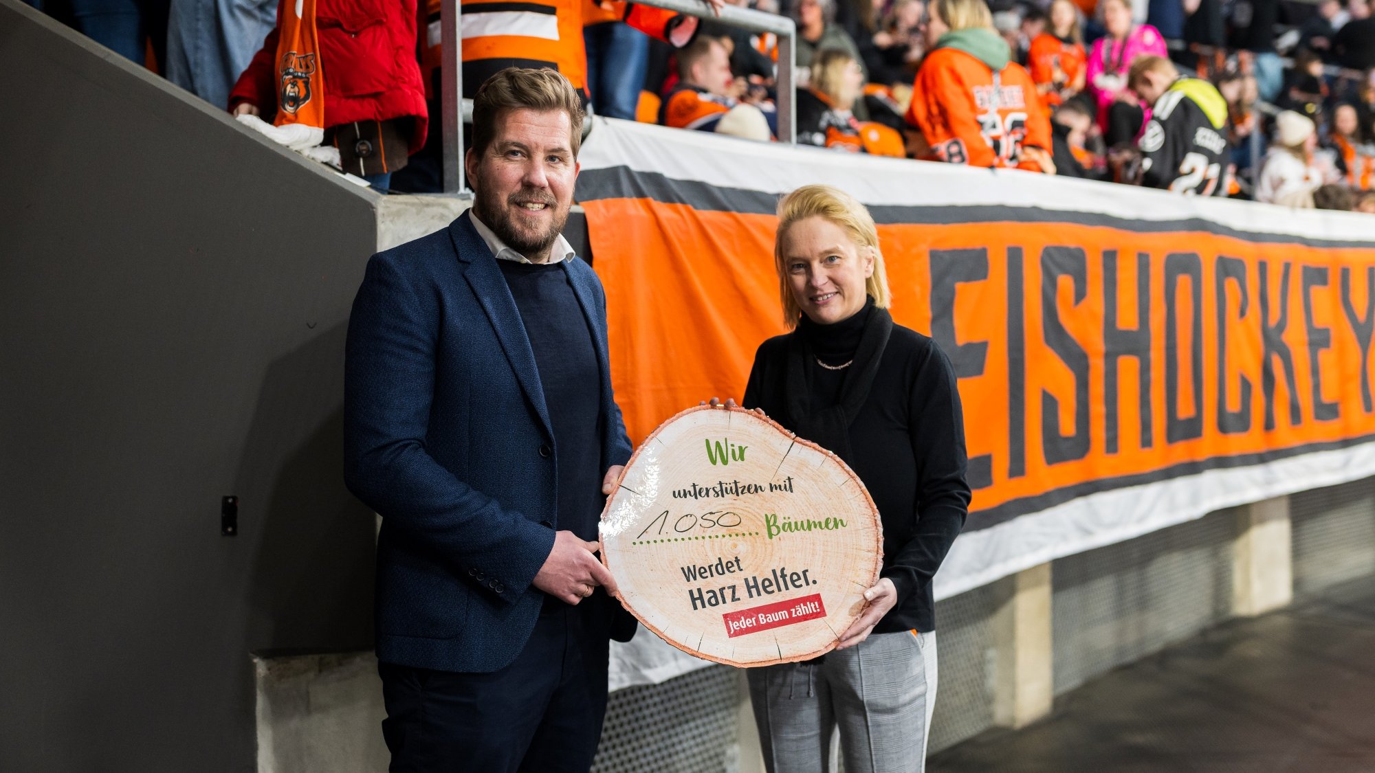 Zwei Personen stehen in einer Sportarena vor einer großen Fahne mit der Aufschrift "Eishockey". Die Person links trägt einen blauen Anzug, die Person rechts einen schwarzen Pullover. Beide halten gemeinsam ein Schild in Holzoptik, auf dem steht: "Wir unterstützen mit 1.050 Bäumen. Werdet Harz Helfer. Jeder Baum zählt!" Im Hintergrund sind Zuschauer in orangefarbenen Trikots zu sehen.