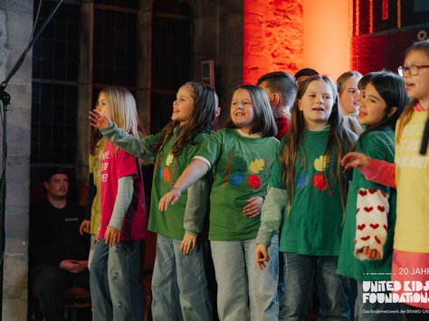 Eine Gruppe von Kindern steht gemeinsam auf einer Bühne und singt. Sie tragen bunte T-Shirts mit verschiedenen Aufdrucken und schauen fröhlich drein. Im Hintergrund sind einige Erwachsene zu sehen, die sich im Schatten aufhalten. Der Raum ist in warmen Tönen beleuchtet, und ein Mikrofonständer ist links im Bild zu erkennen.