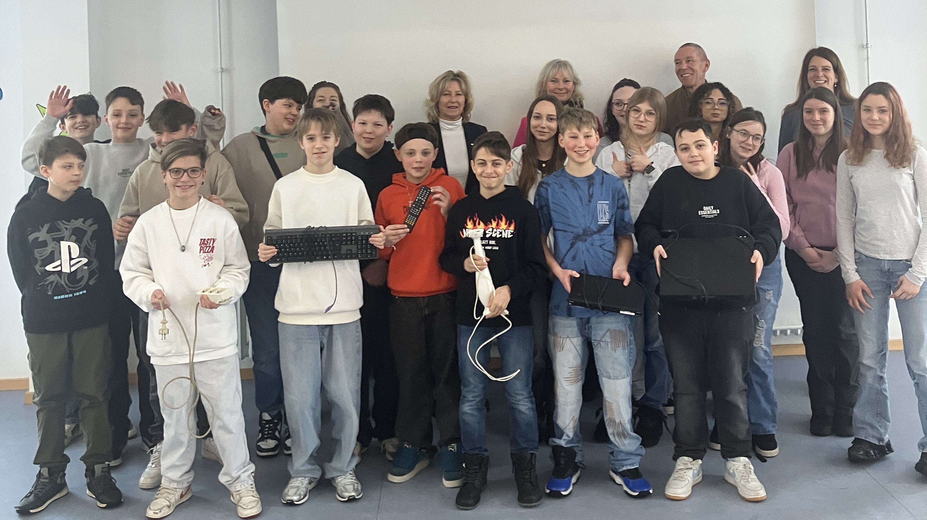 Eine Gruppe von Schülern und Erwachsenen posiert in einem hellen Raum vor einer leeren Projektionsfläche. Einige der Schüler halten Computertastaturen und Kabel in den Händen.