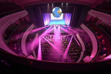Eine Theaterbühne aus der Vogelperspektive. In der Mitte des Theatersaals hängen Scheinwerfer, die pinkfarbenes Licht aussenden, das auf das Publikum gerichtet ist, welches in den Sitzreihen sitzt. Über der Bühne ist ein großer Bildschirm mit dem Bild der Erde und der Jahreszahl 2024 zu sehen. Die Architektur des Saals ist geschwungen, mit mehreren Sitzebenen an den Seiten.
